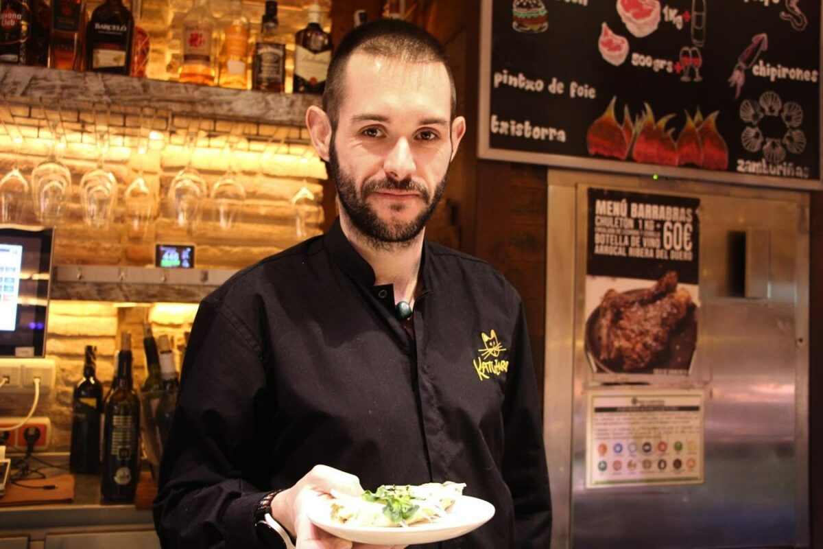 Iker Pérez, camarero del bar Katuzarra, en un plano medio, sostiene un plato con un pincho mientras sonríe a cámara.